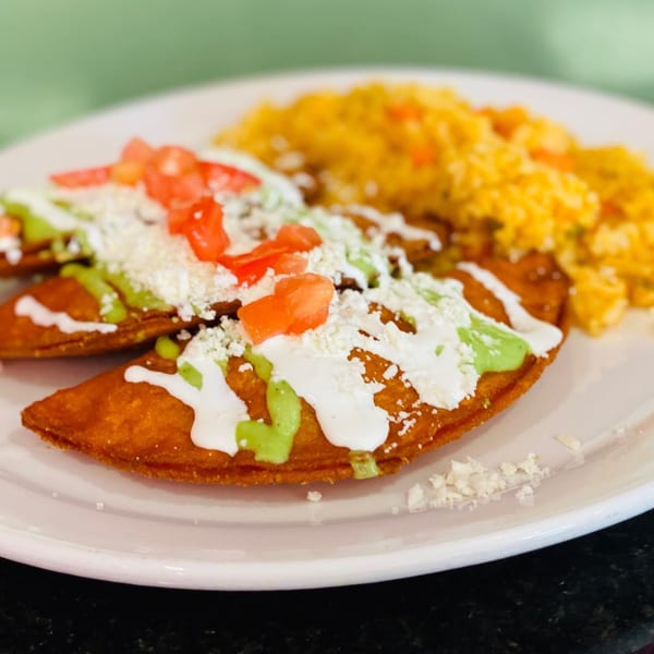 Two golden-brown empanadas topped with white cream, green sauce, tomatoes, and cheese, served with yellow rice.