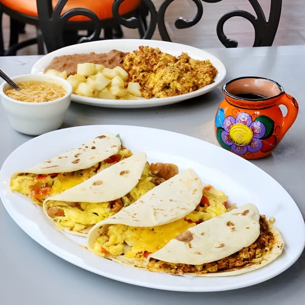 Mexican breakfast with tacos, scrambled eggs, refried beans, potatoes, soup, and a colorful mug.