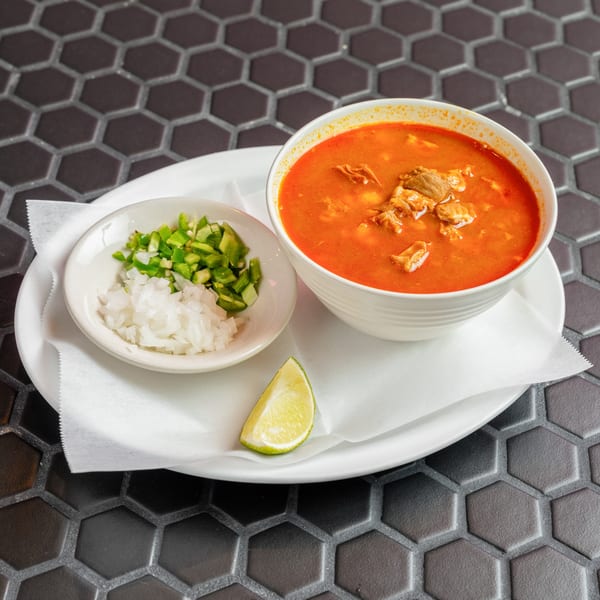 A bowl of Mexico Lindo's red menudo soup with a side of chopped onions, jalapeños, and lime.