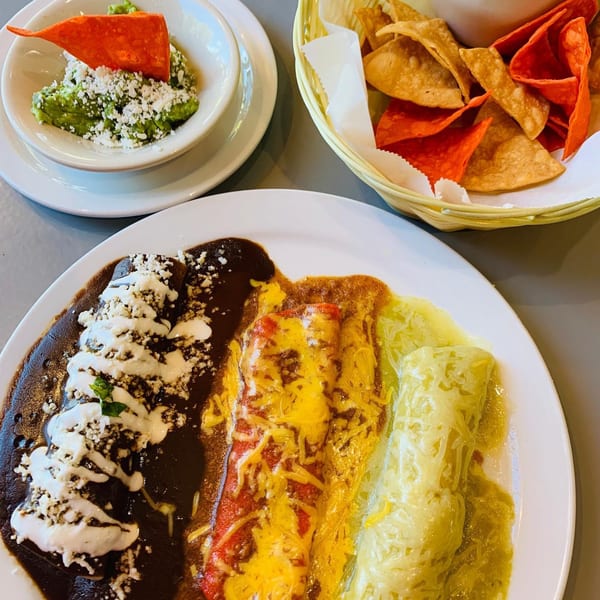A plate of enchiladas with three different sauces, guacamole, and a basket of tortilla chips.