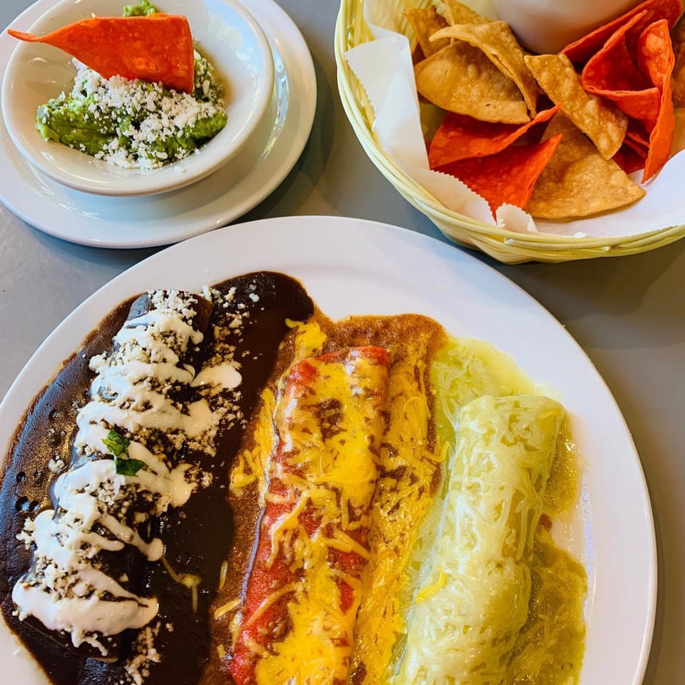 A plate of enchiladas with three different sauces, guacamole, and a basket of tortilla chips.